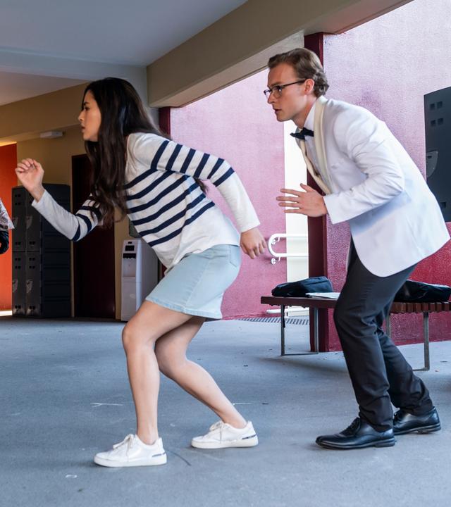 Kyra und Peter gehen in geduckter Haltung und mit angespannter Miene hintereinander her. Aus dem Hintergrund läuft eine Gestalt in einem altmodischen Taucheranzug auf sie zu. Alle drei befinden sich in einem offenen Vorraum einer Schule mit Schließschränken an den Wänden.