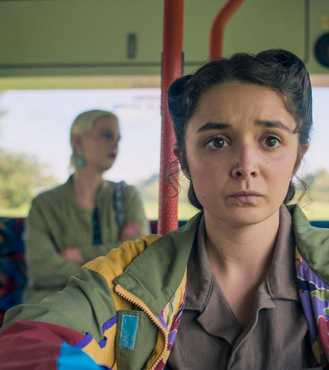 Addie sitzt mit unglücklichem Blick im Bus und schaut gebannt nach vorne. Im Hintergrund, einige Reihen hinter ihr, auf der letzten Bank des Busses sitzt Bonnie. Sie hält die Arme verschränkt vor ihren Körper und schaut traurig blickend nach draußen.