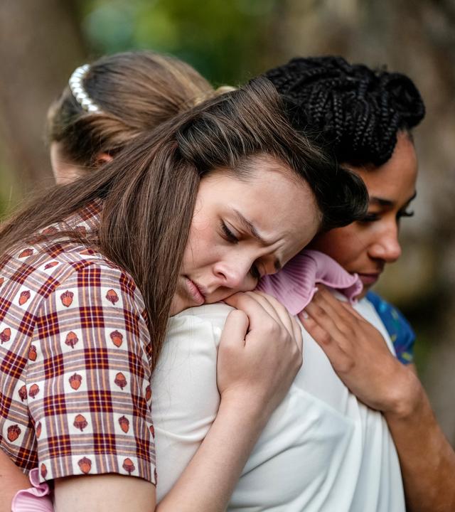 Jenna, Emily und Audrey befinden sich draußen und stehen eng umschlungen und traurig blickend beieinander.