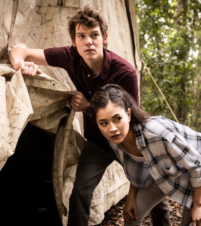 Darra steht neben einem Zelt und hält eine Zeltplane hoch, damit Kyra, die etwas gebeugt neben ihm steht, in das Zelt gehen kann. Beide schauen aufmerksam in eine Richtung.