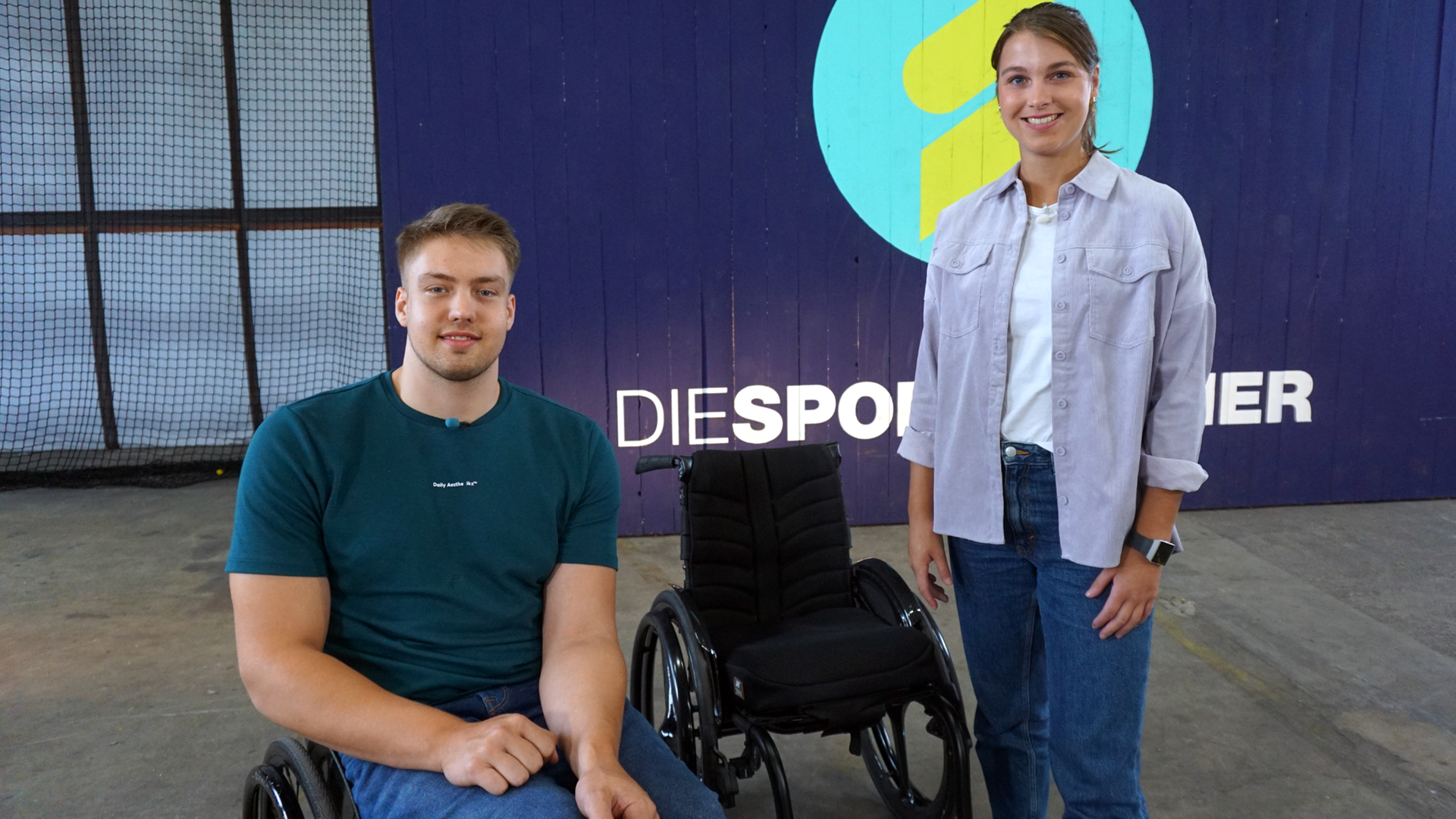 Die Sportmacher: Gast Alex Budde im Rollstuhl sitzend. Rechts daneben Laura Knöll. Mittig von beiden steht ein schwarzer Rollstuhl. Im Hintergrund zu sehen das Sportmacher-Logo.