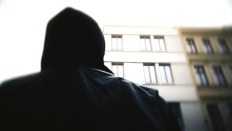 "Stalking - Die unterschätzte Gefahr": Eine dunkel gekleidete Person mit Kapuze, die nur von hinten zu sehen ist, steht vor einem Haus.