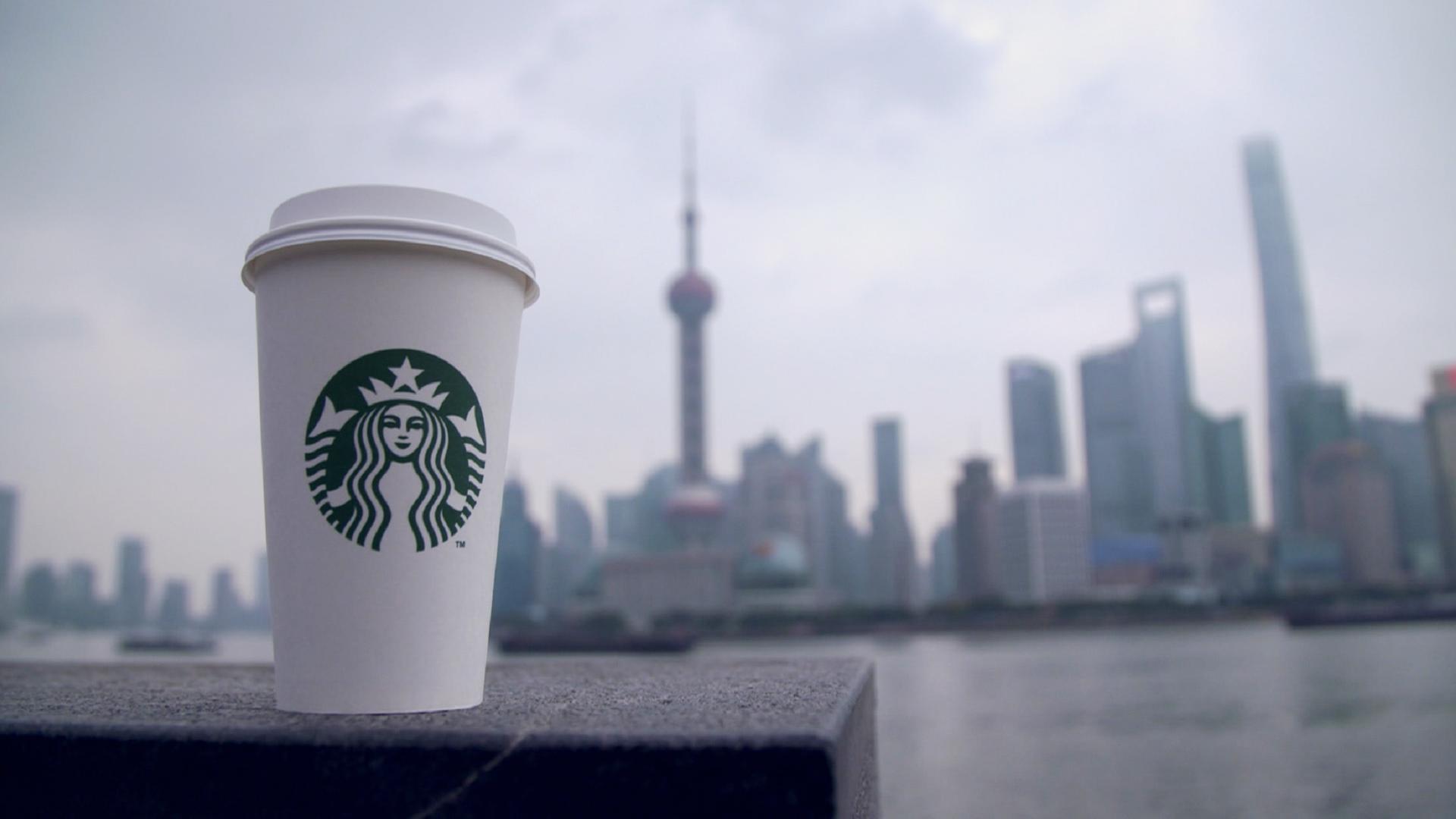 "Starbucks ungefiltert: Die bittere Wahrheit hinter dem Erfolg": Ein Starbucks-Becher steht auf einer Mauer. Im Hintergrund sind verschwommen ein Fluss und die Skyline einer Stadt zu sehen.