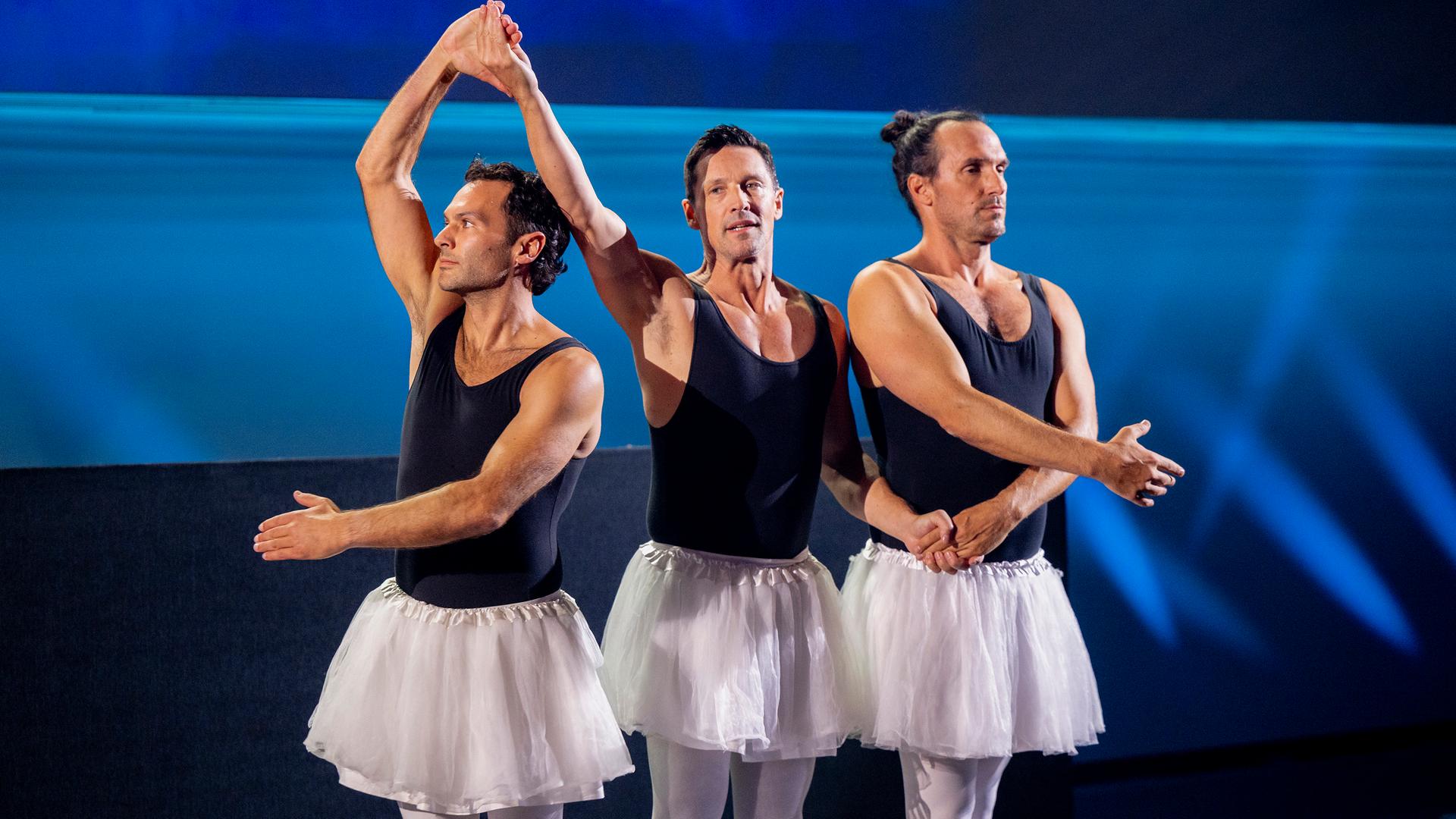 Drei Männer stehen in der Mitte der Bühne und haben Ballettröcke an. Sie halten sich an den Händen. Der Hintergrund ist blau beleuchtet.