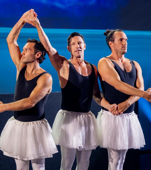 Drei Männer stehen in der Mitte der Bühne und haben Ballettröcke an. Sie halten sich an den Händen. Der Hintergrund ist blau beleuchtet.