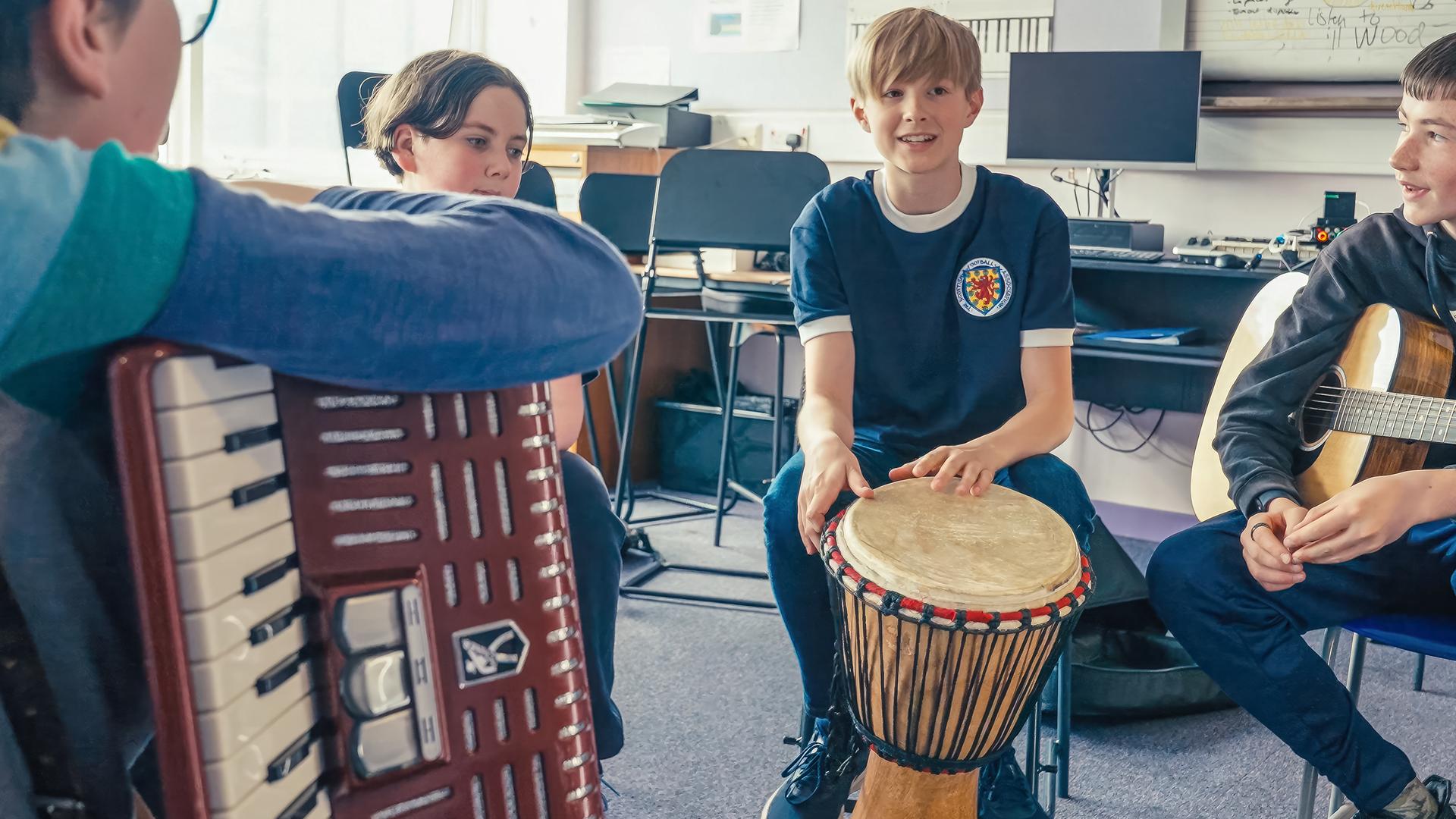 Calum sitzt auf einem Stuhl in einem Klassenzimmer. Er hält eine Trommel in der Hand. Um ihn herum sitzen weitere Schüler, die alle unterschiedliche Musikinstrumente halten.