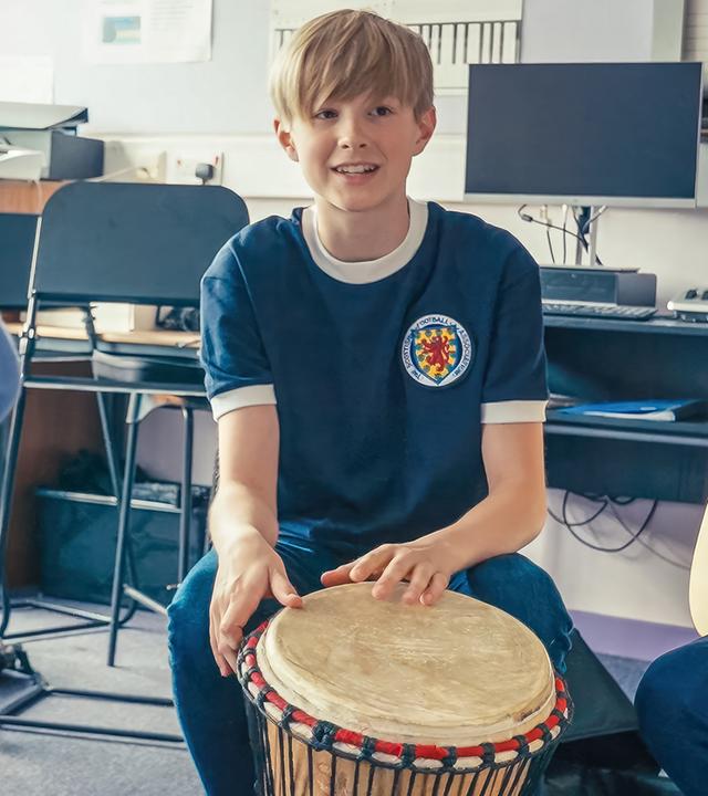 Calum sitzt auf einem Stuhl in einem Klassenzimmer. Er hält eine Trommel in der Hand. Um ihn herum sitzen weitere Schüler, die alle unterschiedliche Musikinstrumente halten.