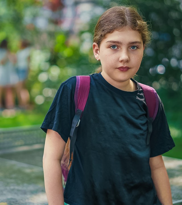 Lya (11) trägt ein schwarzes T-Shirt und einen lila Rucksack. Sie schaut ernst in die Kamera.