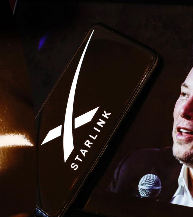 Ein Handy, auf dessen Bildschirm ein weißes X und das Wort "Starlink" zu sehen sind. Daneben ist das Gesicht eines Mannes (Elon Musk) mit dunklen Haaren und in einem Anzug zu sehen, der ein Mikrofon in der Hand hält.