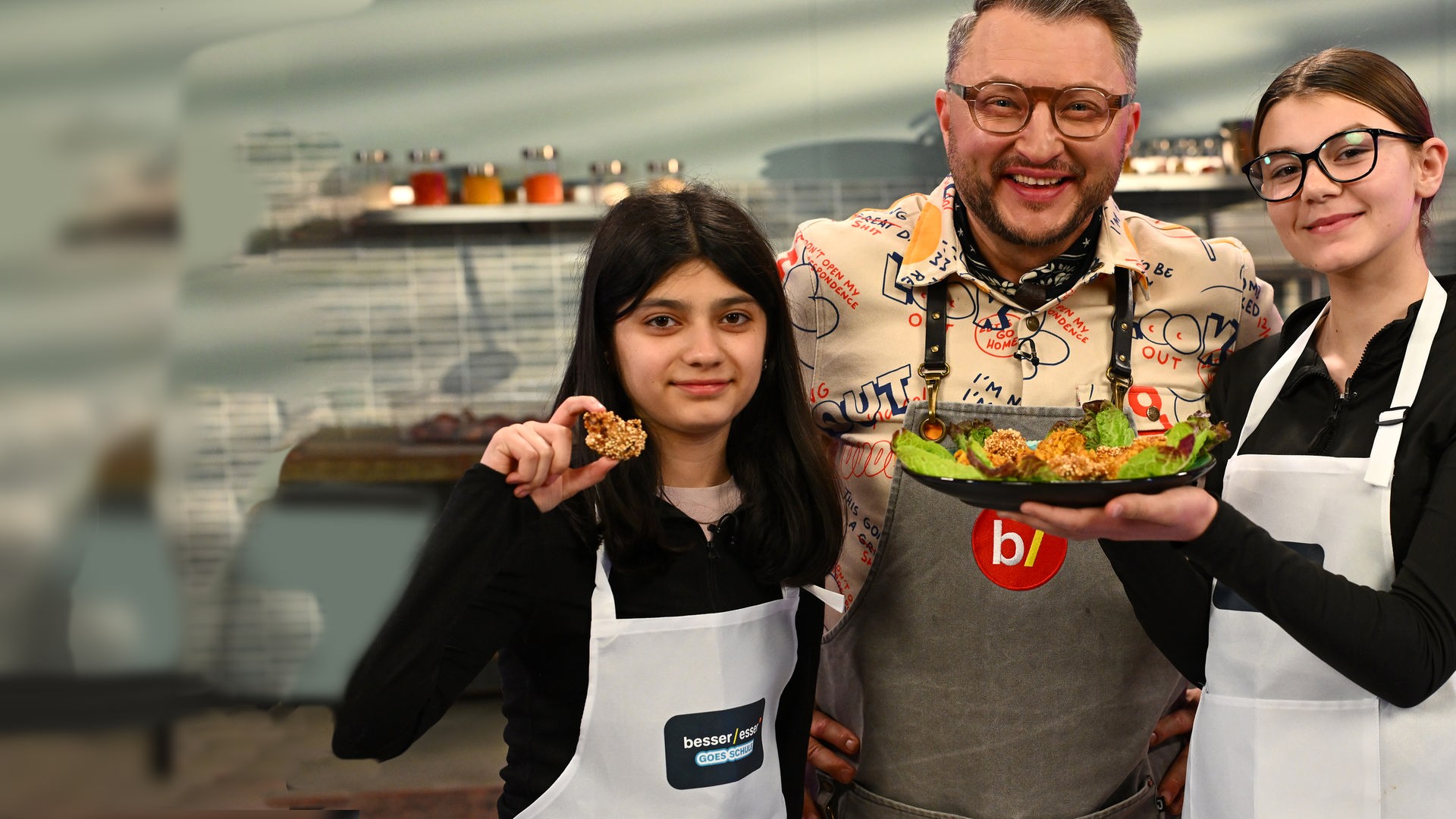 "besseresser Kids": Bibi (l.) und Lucy (r.) zeigen lächelnd ein Tablett voller selbstgemachter Chicken Nuggets mit verschiedenen knusprigen Panaden. Sebastian Lege (m.) blickt zwischen den beiden hervor. Die drei tragen Schürzen und stehen vor der Küchenzeile des Besseresser-Küchenstudios.