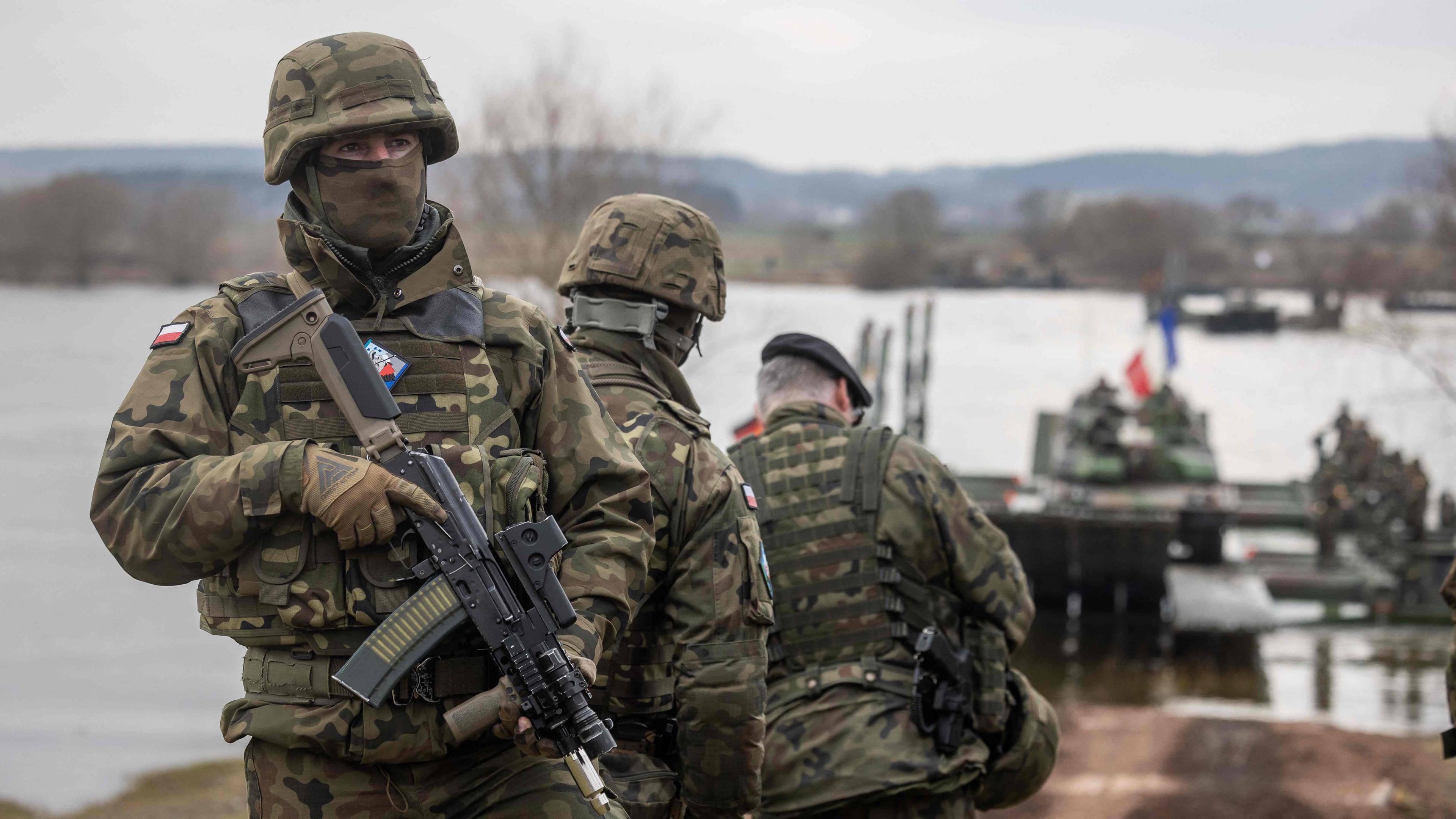 Es sind drei Soldaten zu sehen. Sie stehen vor einem Fluss und im Hintergrund sind Panzer zu sehen.