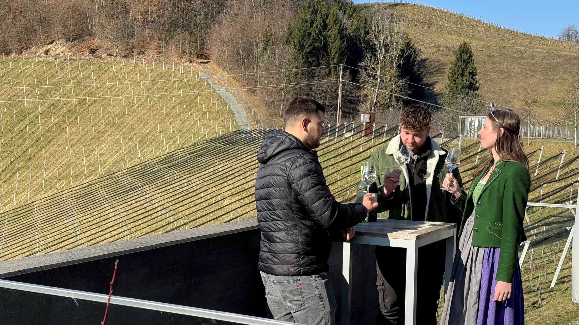 Das Bild zeigt eine ländliche Weinregion mit sanften Hügeln, die mit Weinstöcken bepflanzt sind. Im Vordergrund stehen drei Personen um einen kleinen Tisch. Zwei Männer und eine Frau sind zu sehen. Der erste Mann trägt eine schwarze, gepolsterte Jacke und schaut auf den Tisch, während er mit der linken Hand eine Flasche in der Hand hält. Der zweite Mann hat lockige Haare, trägt eine grüne Jacke und hält ein Weinglas vor sich, während er die Frau ansieht. Die Frau hat lange Haare, trägt eine grüne Jacke und einen langen, grauen Rock mit lila Elementen. Auch sie hält ein Weinglas. Im Hintergrund erkennt man mehrere Reihen von Weinstöcken, die in die Landschaft eingebettet sind. Der Himmel ist klar und die Landschaft wirkt frühlingshaft.