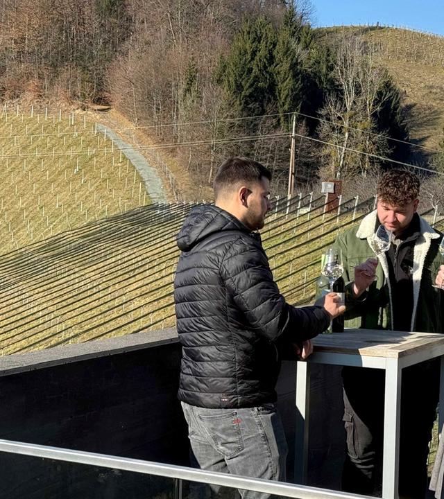 Das Bild zeigt eine ländliche Weinregion mit sanften Hügeln, die mit Weinstöcken bepflanzt sind. Im Vordergrund stehen drei Personen um einen kleinen Tisch. Zwei Männer und eine Frau sind zu sehen. Der erste Mann trägt eine schwarze, gepolsterte Jacke und schaut auf den Tisch, während er mit der linken Hand eine Flasche in der Hand hält. Der zweite Mann hat lockige Haare, trägt eine grüne Jacke und hält ein Weinglas vor sich, während er die Frau ansieht. Die Frau hat lange Haare, trägt eine grüne Jacke und einen langen, grauen Rock mit lila Elementen. Auch sie hält ein Weinglas. Im Hintergrund erkennt man mehrere Reihen von Weinstöcken, die in die Landschaft eingebettet sind. Der Himmel ist klar und die Landschaft wirkt frühlingshaft.