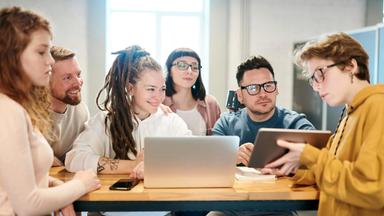 Fünf junge Erwachsene sitzen gemeinsam an einem Tisch in einem hellen Büro und arbeiten im Team. Zwei Personen nutzen Laptops, während eine weitere ein Tablet zeigt und die anderen aufmerksam folgen.