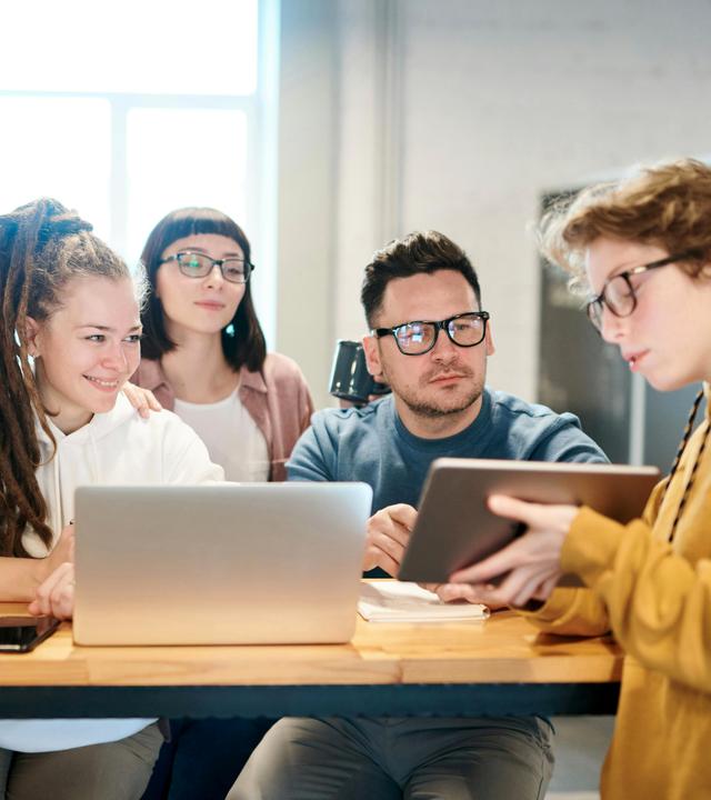 Fünf junge Erwachsene sitzen gemeinsam an einem Tisch in einem hellen Büro und arbeiten im Team. Zwei Personen nutzen Laptops, während eine weitere ein Tablet zeigt und die anderen aufmerksam folgen.