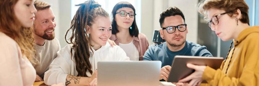 Fünf junge Erwachsene sitzen gemeinsam an einem Tisch in einem hellen Büro und arbeiten im Team. Zwei Personen nutzen Laptops, während eine weitere ein Tablet zeigt und die anderen aufmerksam folgen.