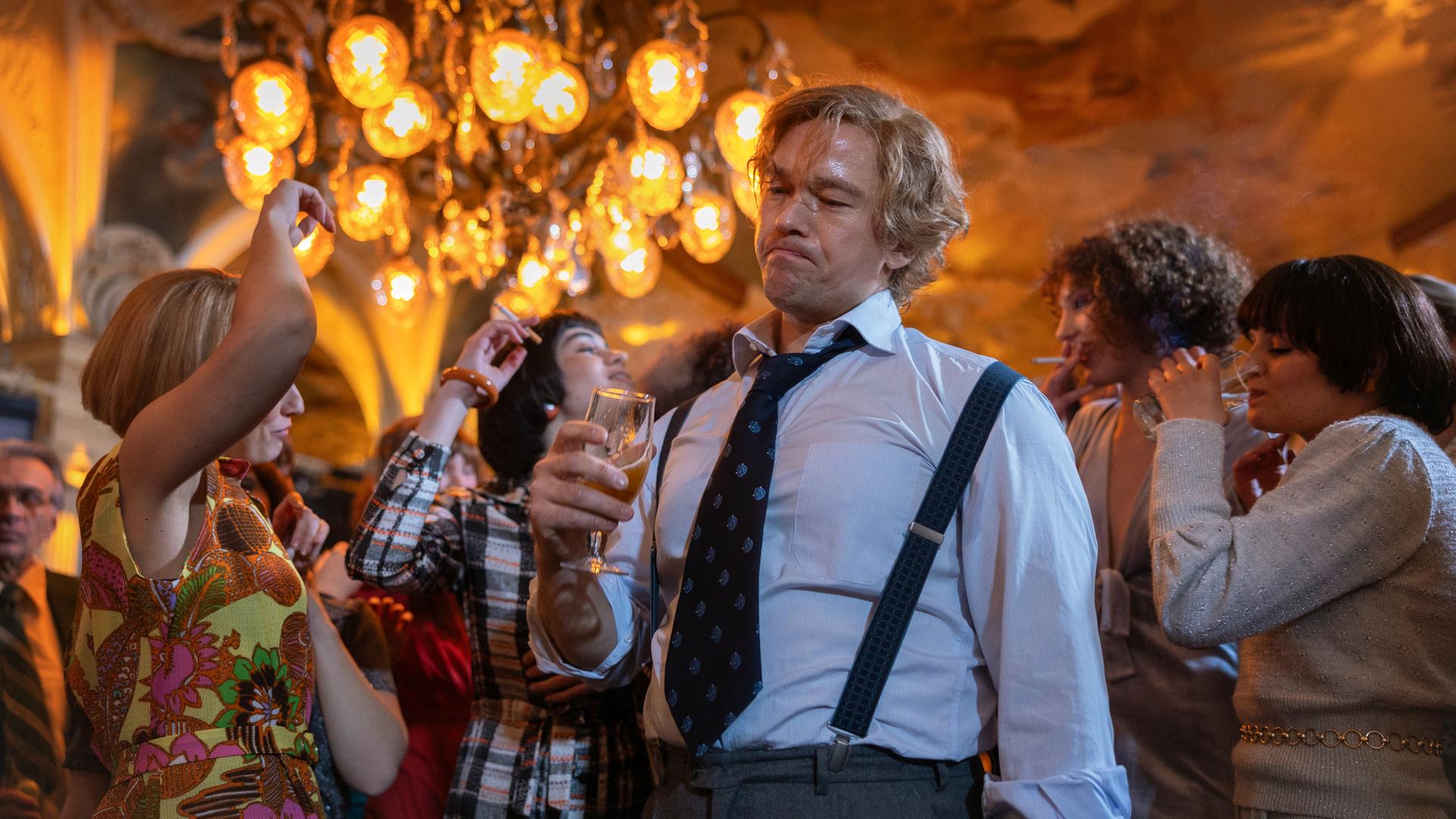 "Stenbeck - Zwischen zwei Welten": Im Büro-Outfit mit Hemd, Krawatte und Hosenträgern steht Jan Stenbeck (Jakob Oftebro) mit einem Bierglas in der Hand unter einem Kronleuchter, umgeben von tanzenden Frauen (Komparsinnen).