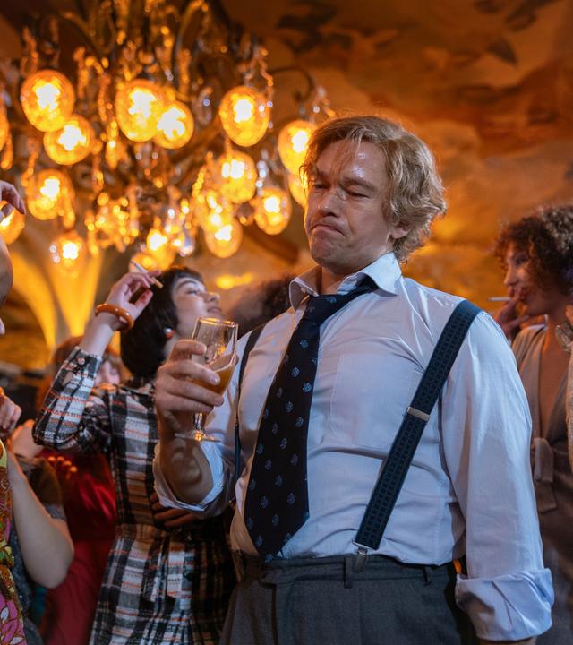 "Stenbeck - Zwischen zwei Welten": Im Büro-Outfit mit Hemd, Krawatte und Hosenträgern steht Jan Stenbeck (Jakob Oftebro) mit einem Bierglas in der Hand unter einem Kronleuchter, umgeben von tanzenden Frauen (Komparsinnen).