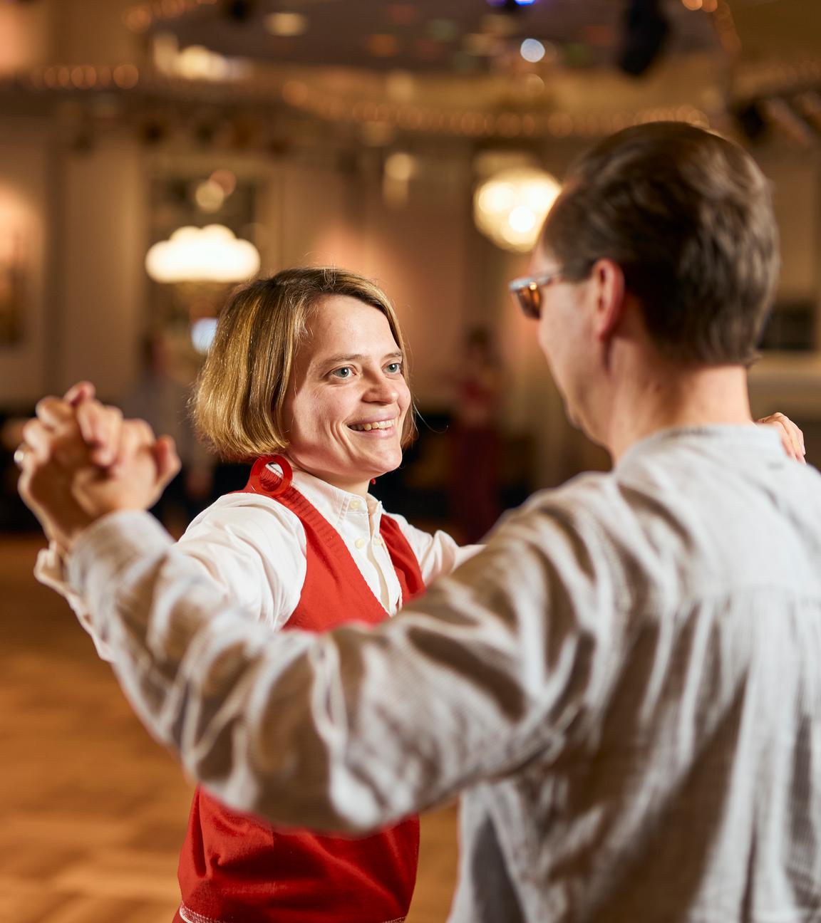 Kathrin und Kristian stehen in einer Standardtanzposition in einem Tanzsaal in goldgelben Farbtönen. Kathrin strahlt ihren Tanzpartner, der mit dem Rücken zur Kamera steht, an.