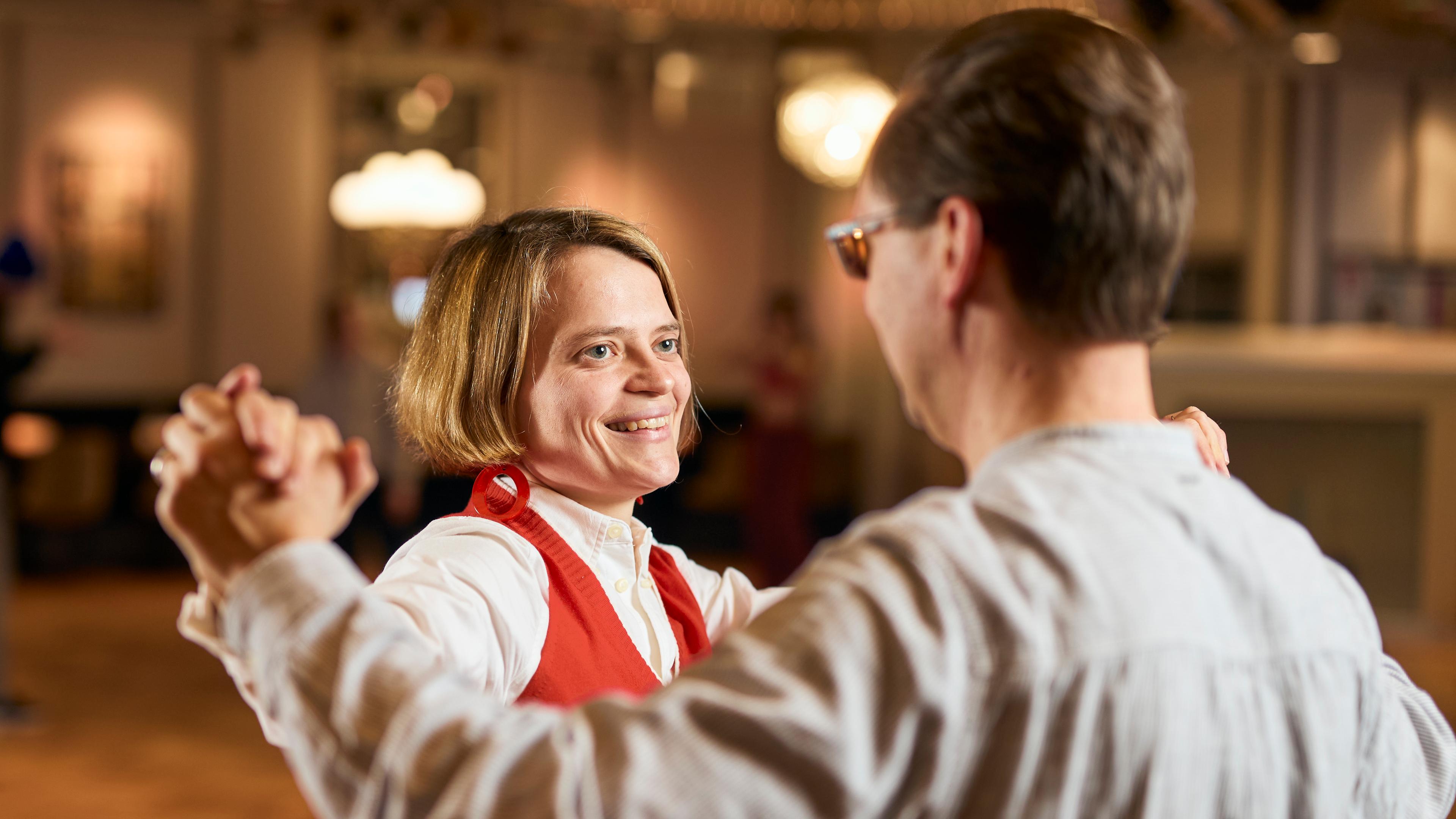Kathrin und Kristian stehen in einer Standardtanzposition in einem Tanzsaal in goldgelben Farbtönen. Kathrin strahlt ihren Tanzpartner, der mit dem Rücken zur Kamera steht, an.