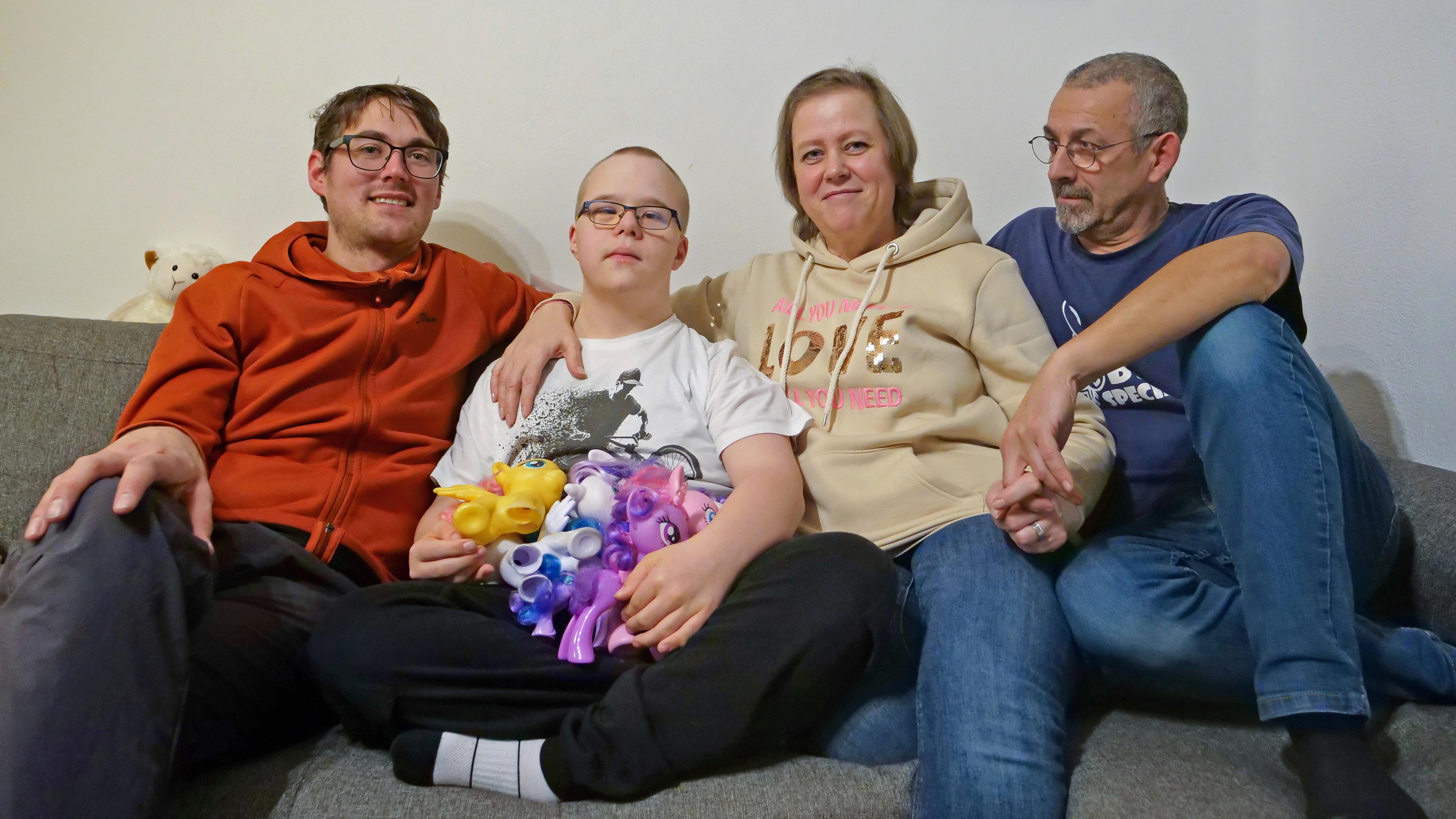 Tobias, ein Mann Mitte Dreißig, sitzt mit Jannes, einem Jungen mit Down-Syndrom, Yvonne, Jannes Mutter und deren Lebenspartner auf einer grauen Sitzecke. Jannes hat Einhörner auf dem Schoß.