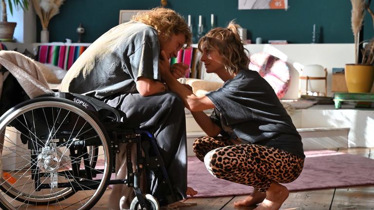 "Sterben für Beginner": Alex (Max Hubacher) sitzt in einem Rollstuhl im Wohnzimmer. Karla (Svenja Jung) hat sich vor ihn gehockt und streichelt seine Wange.