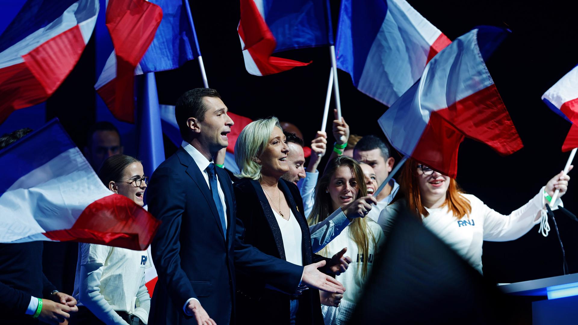 Marine Le Pen und Jordan Bardella auf der Bühne, hinter ihnen stehen, mit der französischen Flagge winkend, junge Menschen.
