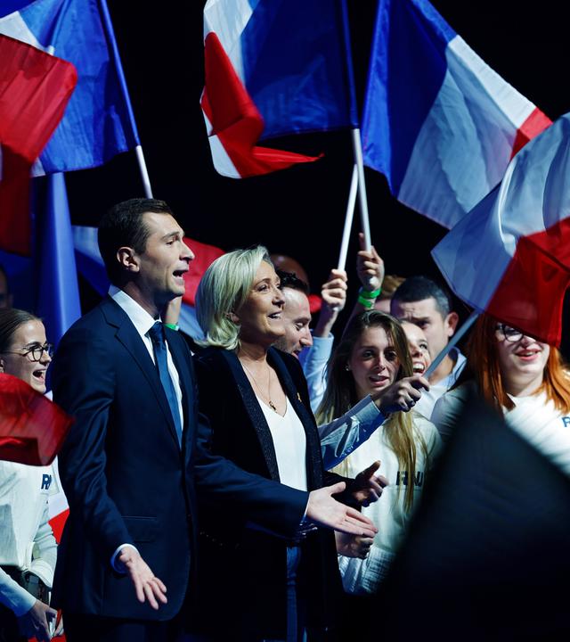 Marine Le Pen und Jordan Bardella auf der Bühne, hinter ihnen stehen, mit der französischen Flagge winkend, junge Menschen.
