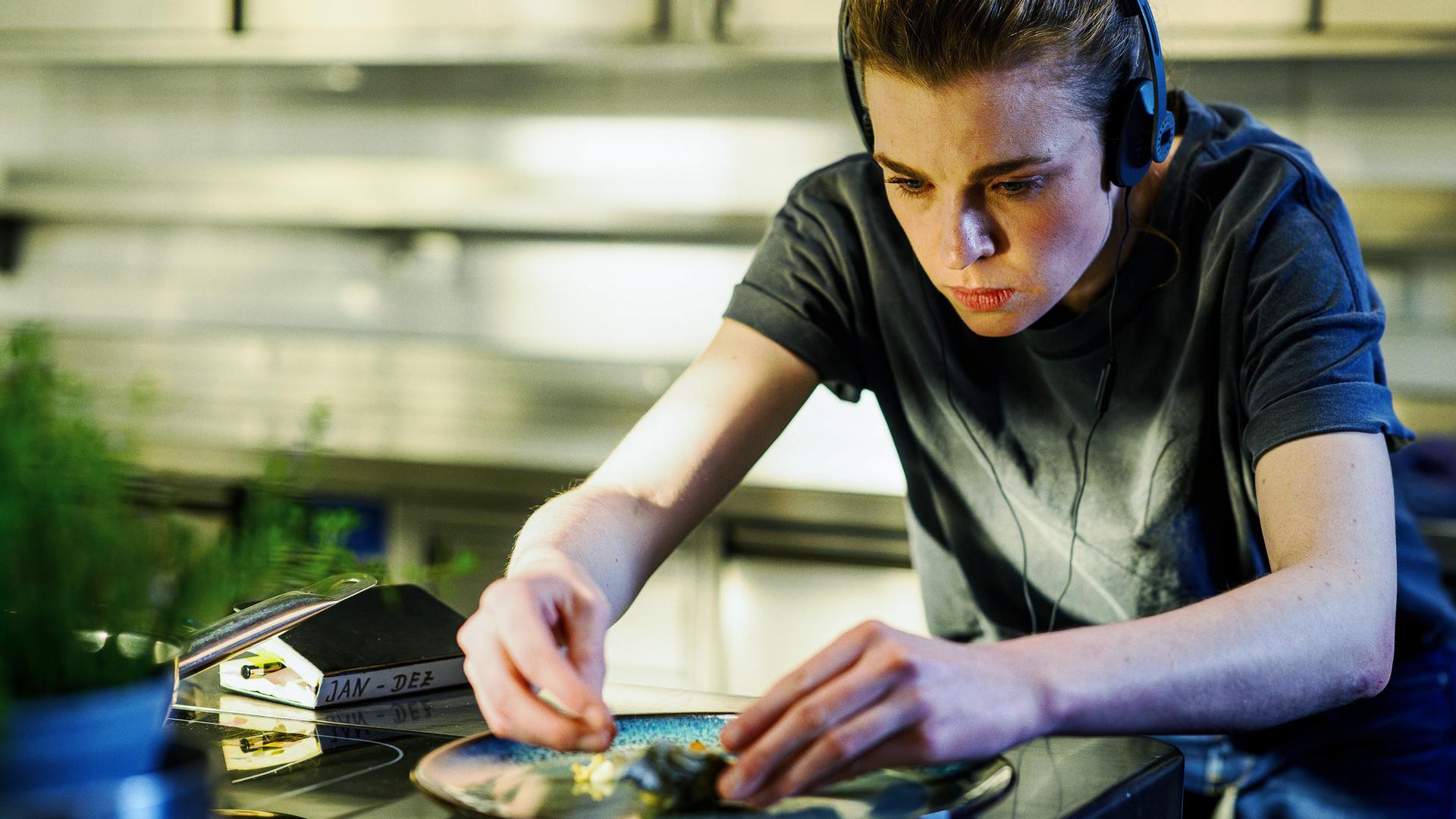 Tanja Möppen (Franziska Ferrari) steht in der Küche und richtet einen Teller mit Speisen an. Neben ihr liegt ein Notizbuch und sie trägt Kopfhörer.