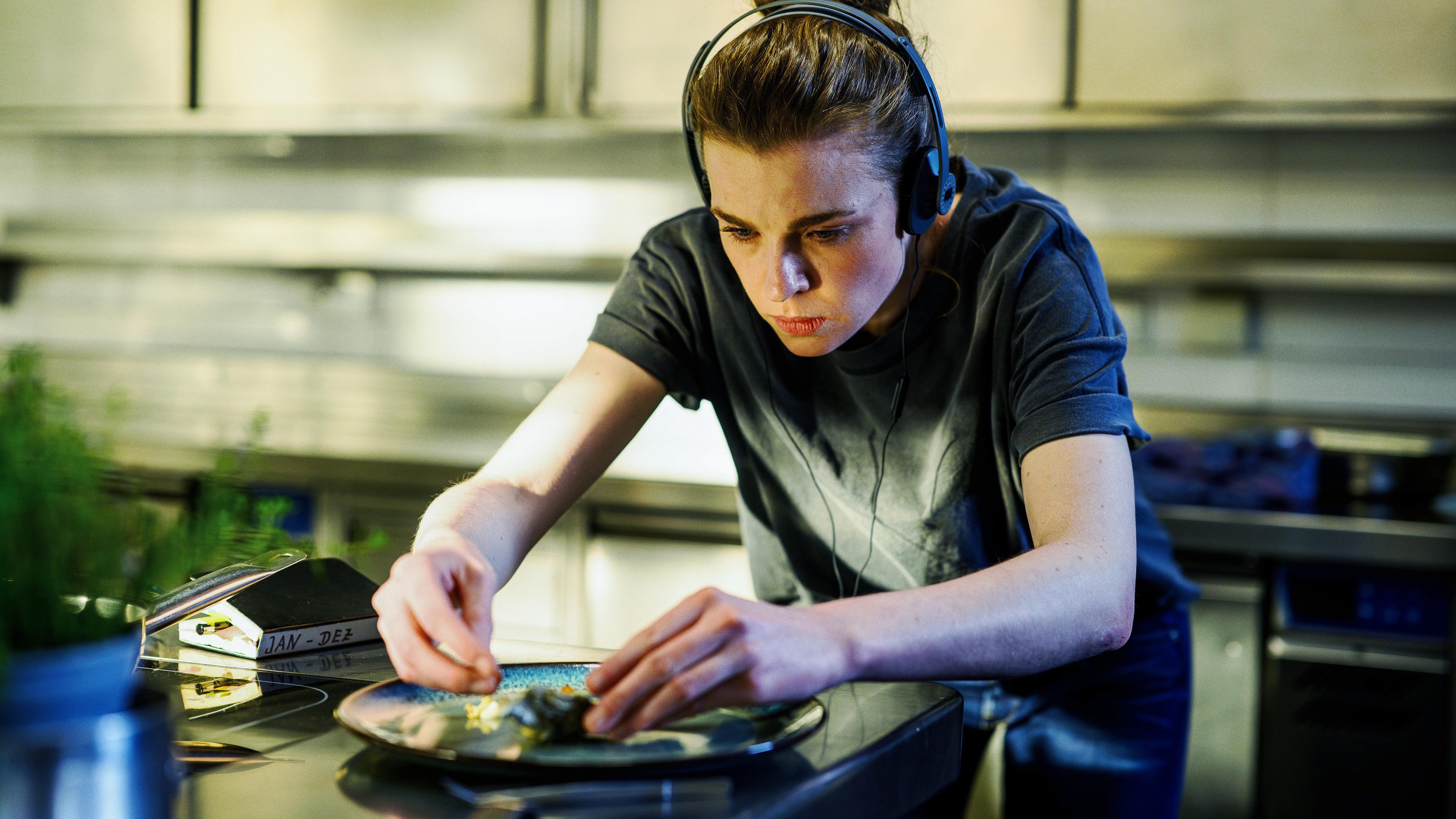 Tanja Möppen (Franziska Ferrari) steht in der Küche und richtet einen Teller mit Speisen an. Neben ihr liegt ein Notizbuch und sie trägt Kopfhörer.