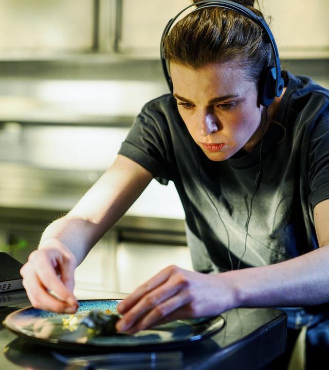 Tanja Möppen (Franziska Ferrari) steht in der Küche und richtet einen Teller mit Speisen an. Neben ihr liegt ein Notizbuch und sie trägt Kopfhörer.