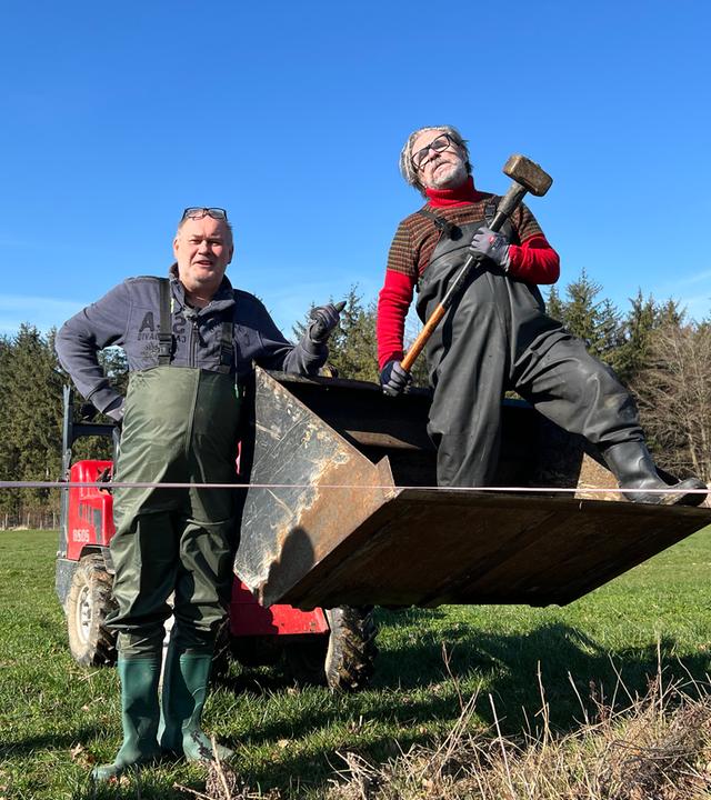 Waldi und sein Freund Ricardo stehen am Zaun einer Wiese. Ricardo steht in einer Baggerschaufel und hält einen Vorschlaghammer.