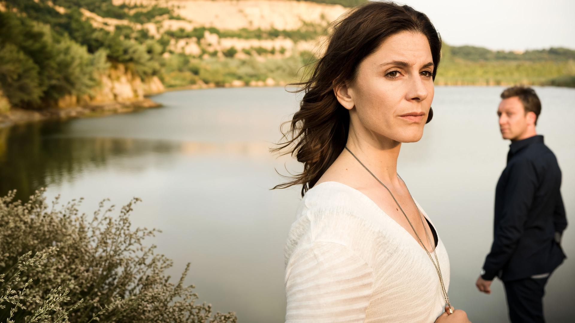 "In Wahrheit - Still ruht der See": Judith Mohn (Christina Hecke) blickt nachdenklich aus dem Bild. Hinter ihr steht Freddy Breyer (Robin Sondermann) und schaut zu Judith hin. Im Hintergrund ist ein See zu sehen.