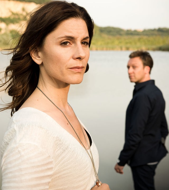 "In Wahrheit - Still ruht der See": Judith Mohn (Christina Hecke) blickt nachdenklich aus dem Bild. Hinter ihr steht Freddy Breyer (Robin Sondermann) und schaut zu Judith hin. Im Hintergrund ist ein See zu sehen.