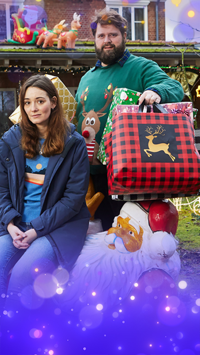 Liane (Maria Ehrich) sitzt auf einer liegenden Weihnachtsmann-Dekoration auf dem Rasen vor einem weihnachtlich geschmückten Haus. Sie schaut amüsiert und blickt in die Kamera. Hinter ihr steht Hinnerk (Antoine Monot), der viele große, bunte Weihachtsgeschenke unter den Armen hält Auch er schaut lachend in die Kamera.