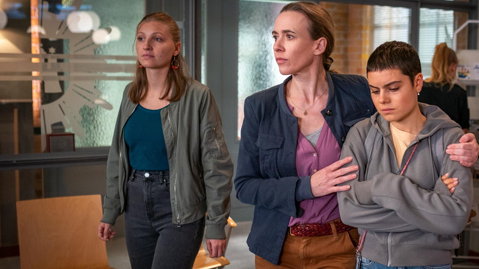  Jenny (Annalisa Weyel), Rita (Kathrin-Marén Enders) und Marlee (Holly Geddert) gehen nebeneinander durch das PK21. Rita hat ihren linken Arm um die Schultern ihrer Tochter gelegt. Marlee hat die Arme vor dem Oberkörper verschränkt und schaut nachdenklich zu Boden.