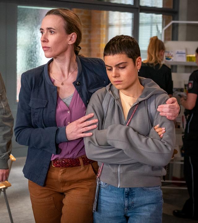  Jenny (Annalisa Weyel), Rita (Kathrin-Marén Enders) und Marlee (Holly Geddert) gehen nebeneinander durch das PK21. Rita hat ihren linken Arm um die Schultern ihrer Tochter gelegt. Marlee hat die Arme vor dem Oberkörper verschränkt und schaut nachdenklich zu Boden.