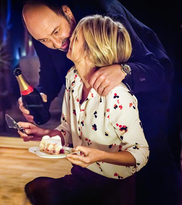 Im gedämpften Licht einer Wohnzimmerlampe beugt sich Thomas Kertez (Kad Merad) mit einer Flasche Sekt in der Hand zu Alice (Laurence Arné), die gerade ein Stück Kuchen auf einem Teller in der Hand hält.