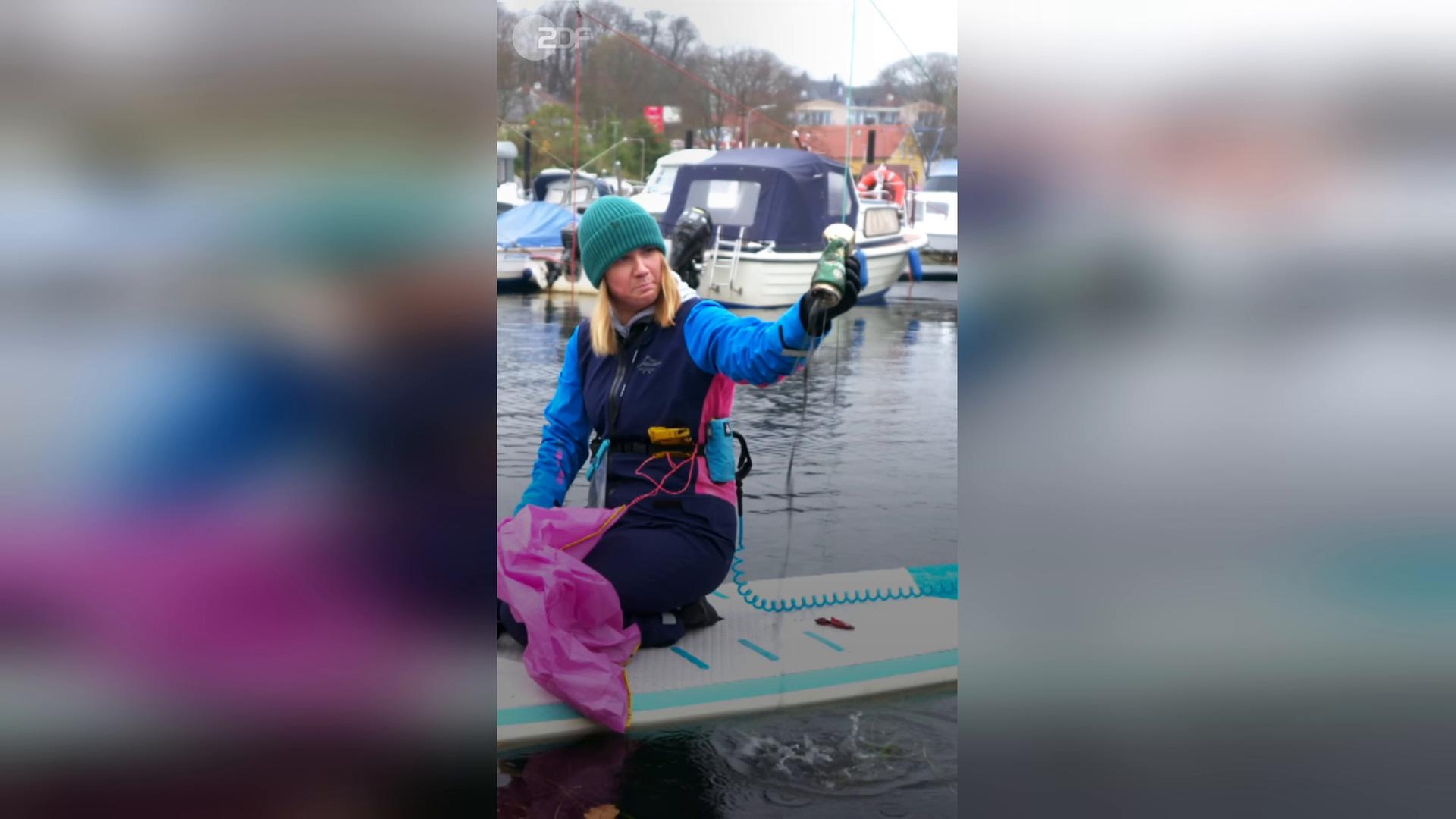 Louisa Dellert kniet im Hafen auf einem Standup-Paddelboard, eine Bierdose in der Hand. 
