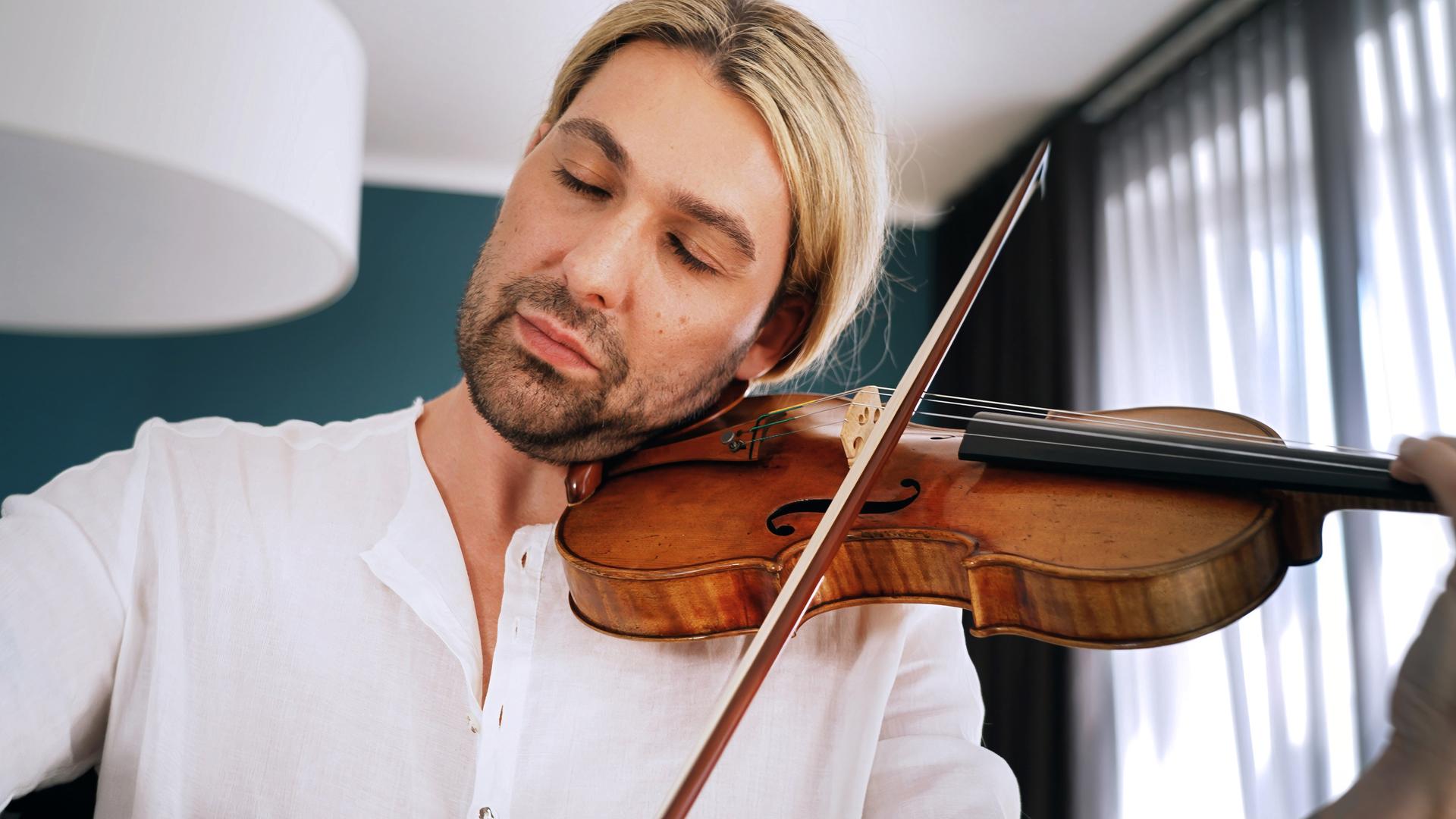 David Garret sitzt in einem Sessel und hält seine Stradivari in seiner Hand und auf seinem Schoß. Er trägt ein lockeres Leinenhemd, auf dem Arm ist deutlich eine Tätowierung sichtbar. Er spricht.
