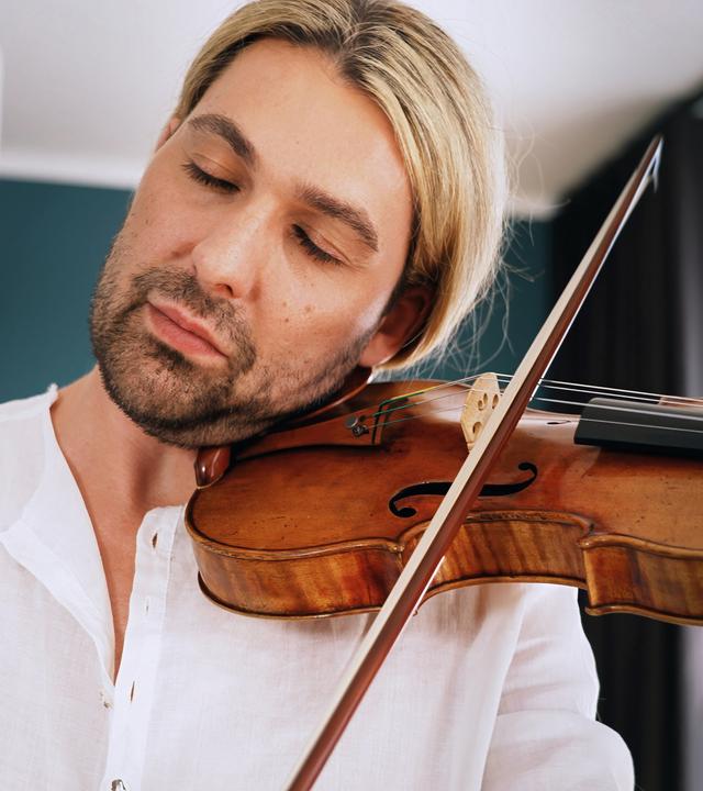 David Garret sitzt in einem Sessel und hält seine Stradivari in seiner Hand und auf seinem Schoß. Er trägt ein lockeres Leinenhemd, auf dem Arm ist deutlich eine Tätowierung sichtbar. Er spricht.