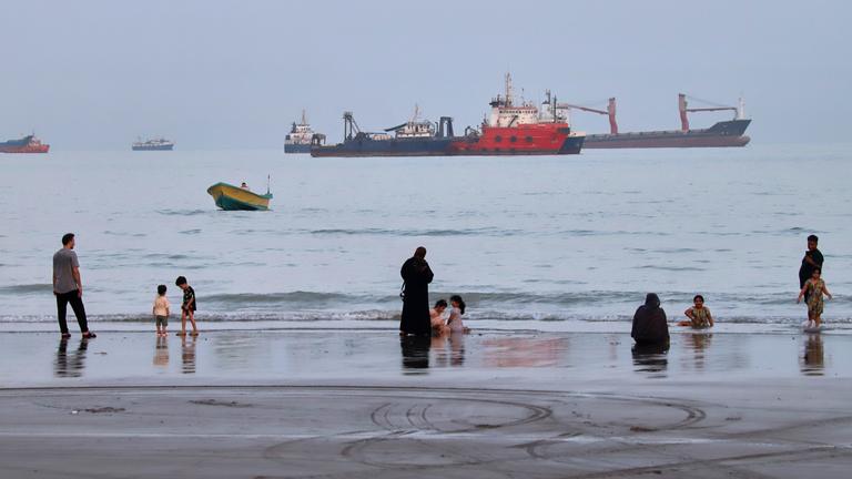 Menschen verbringen Zeit am Strand, während vor Bandar Abbas im Iran in der Straße von Hormus Handelsschiffe vor Anker liegen