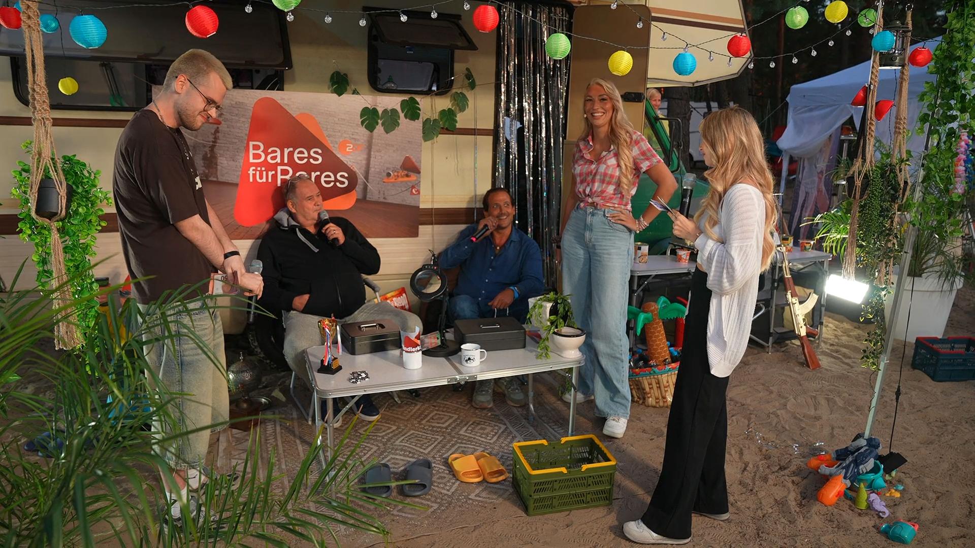 "Streamergarten - Folge 1: Bares für Rares - Streamer-Edition": Bares für Rares - Streamer-Edition": Die Streamer JenNyan (rechts) und Faister (links) stehen mit Host Nova (2.v.r.) in einer Camping-Kulisse. Im Hintergrund ein Campingmobil, vor dem zwei weitere Gäste sitzen.