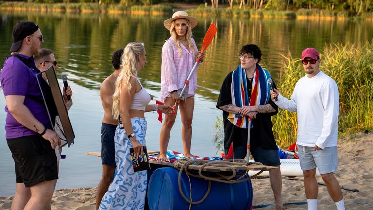 "Streamergarten - Folge 2: Das ultimative Floß-Duell": Milschbaum steht mit einem Paddel in der Hand auf dem aus Tonnen und Brettern gefertigten Floß am Ufer des Sees. Niekbeats, die Wolter Twins und Starletnova stehen um sie herum.
