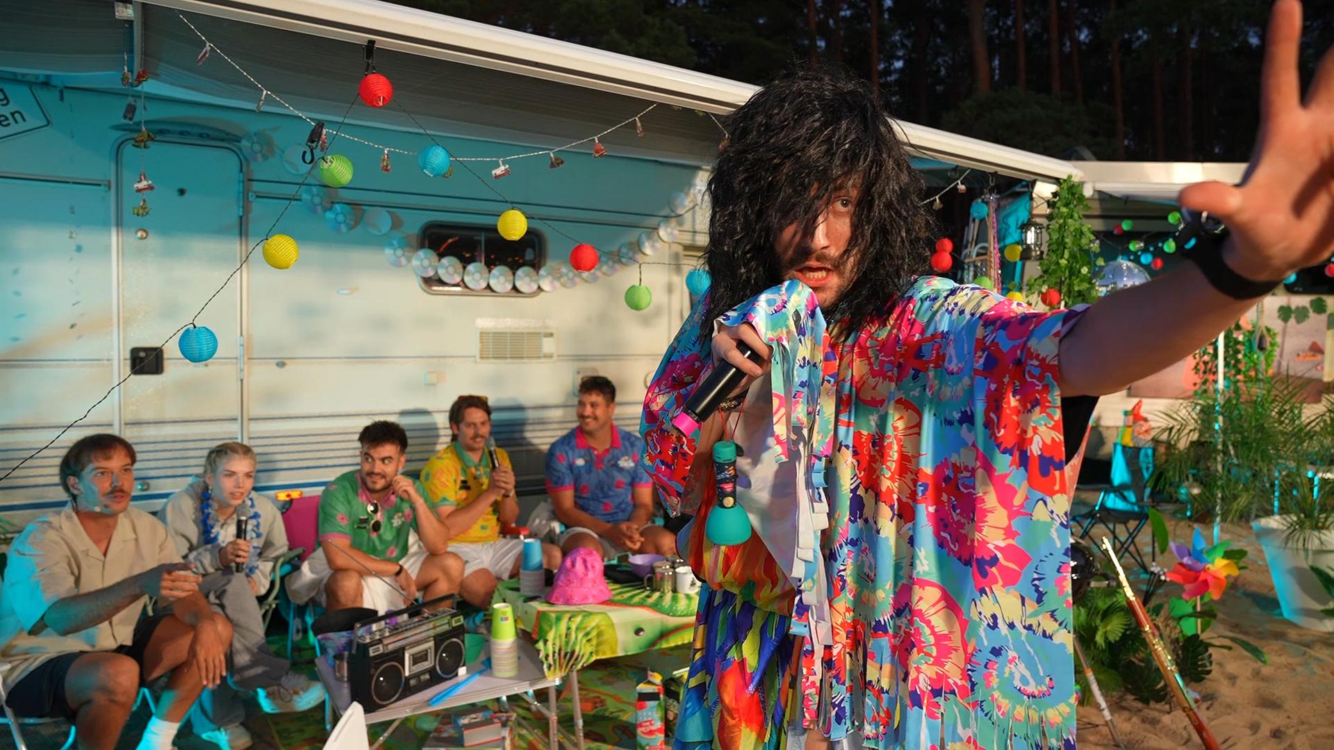 "Streamergarten - Folge 1: Der Sternecoach sucht Liebe": Eine männliche Gestalt mit langen Haaren und mit einem bunten Gewand bekleidet (r.) hält in einer Hand ein Mikrofon, während die andere Hand zu Beschwörung erhoben ist. Im Hintergrund sitzen mehrere Gäste vor einem Campermobil und beobachten gemeinsam mit Host Aditotoro (l.) die Gestalt.