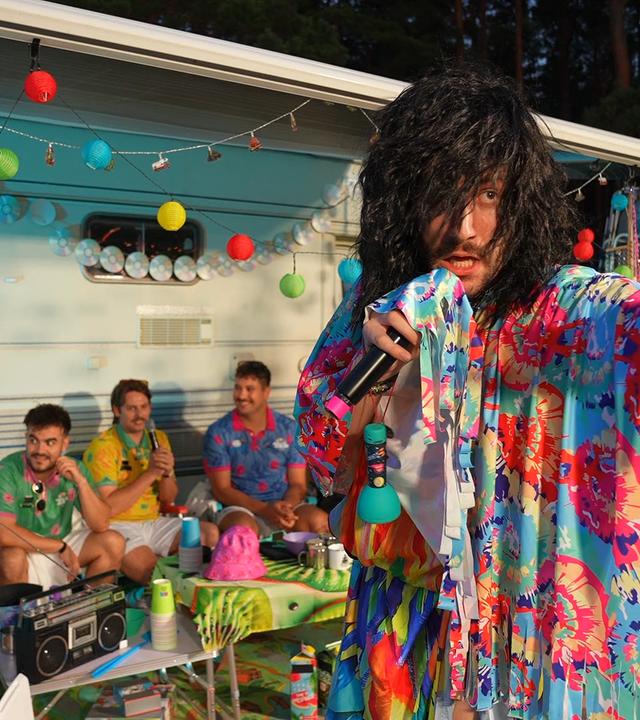 "Streamergarten - Folge 1: Der Sternecoach sucht Liebe": Eine männliche Gestalt mit langen Haaren und mit einem bunten Gewand bekleidet (r.) hält in einer Hand ein Mikrofon, während die andere Hand zu Beschwörung erhoben ist. Im Hintergrund sitzen mehrere Gäste vor einem Campermobil und beobachten gemeinsam mit Host Aditotoro (l.) die Gestalt.