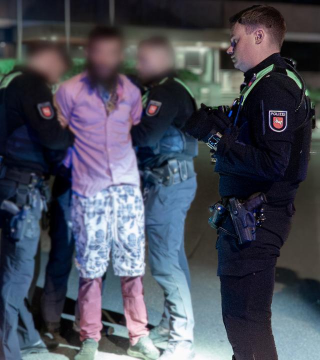 Ein unkenntlich gemachter Mann wird von zwei Polizisten festgehalten, daneben ein weiterer Polizist, der sich etwas notiert. 
