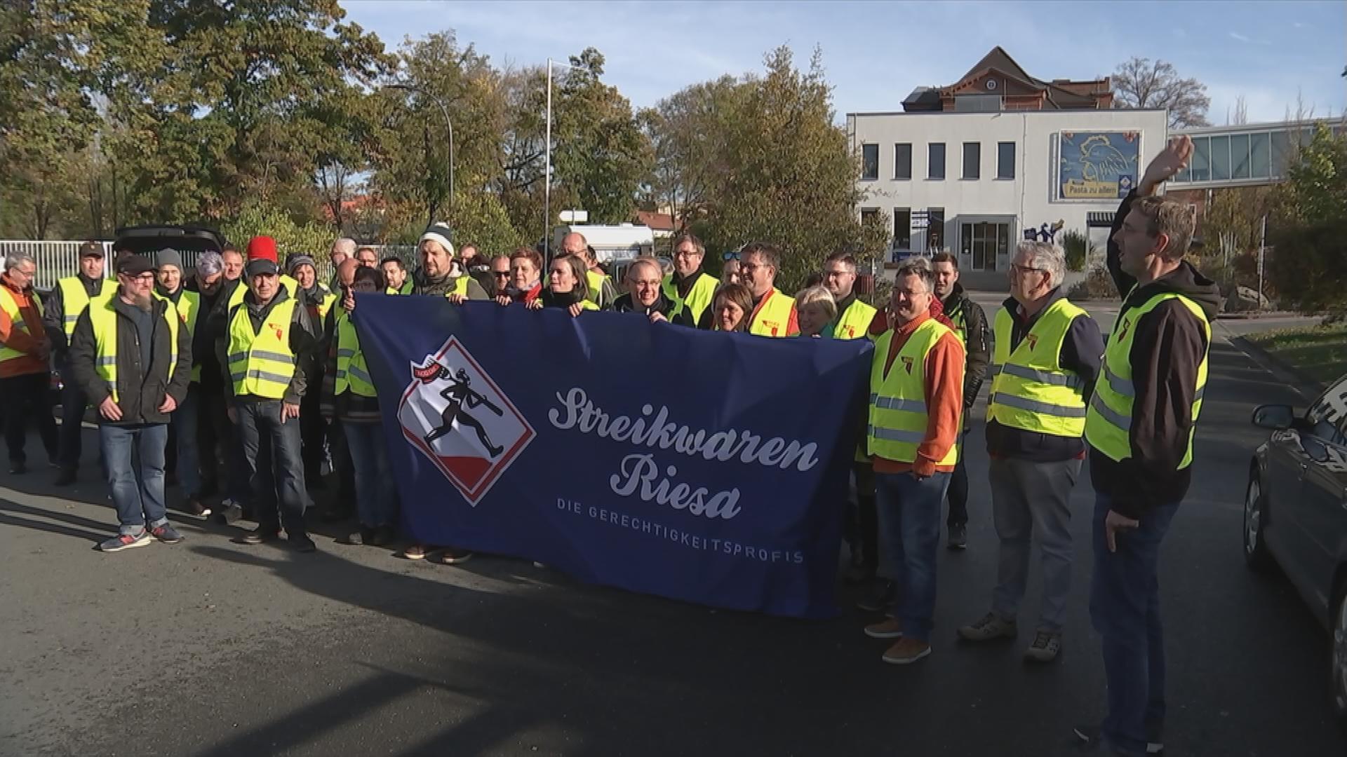 Arbeiter der Firma Riesa Teigwaren mit gelben Warnwesten. Sie halten ein Plakat mit der Aufschrift "Streikwaren Riesa".