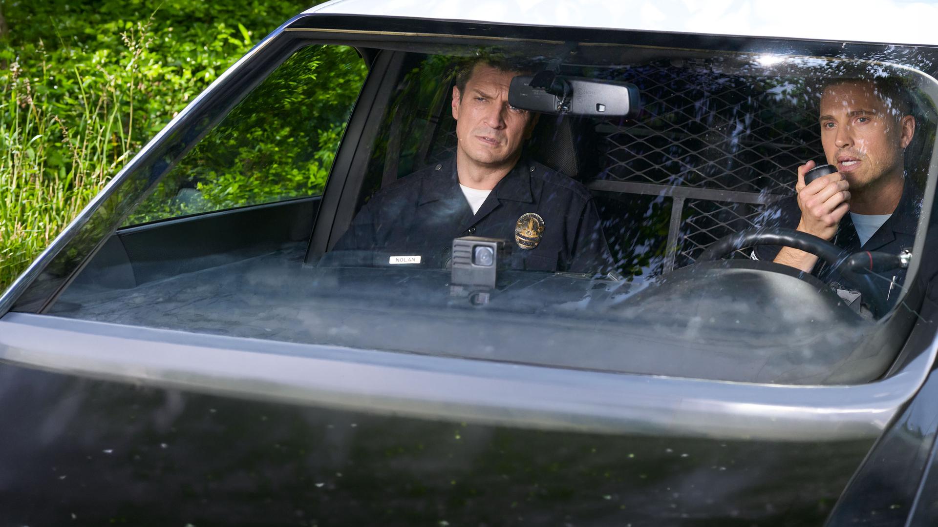 "The Rookie - Stresstest": Nolan (Nathan Fillion) und Bradford (Eric Winter) sitzen nebeneinander in einem Streifenwagen. Während Nolan konzentriert nach vorne schaut, hält Bradford das Funkgerät in seiner Hand.