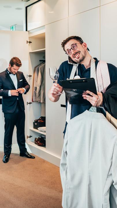 Mann mit Klemmbrett (vorne rechts) und einem Sektglas in der Hand sowie Anzügen auf dem Arm. Ein Handy ist zwischen Ohr und Schultern geklemmt. Weitere Mann im Bildhintergrund links.